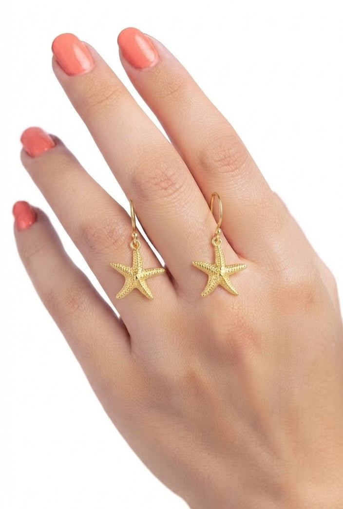 Pendientes estrella de mar dorados sostenidos en mano con uñas coral